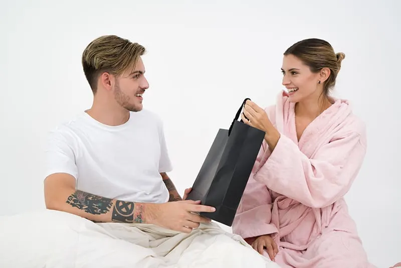 ein lächelnder Mann, der einer Frau eine Tasche mit einem Geschenk gibt, während er auf dem Bett sitzt