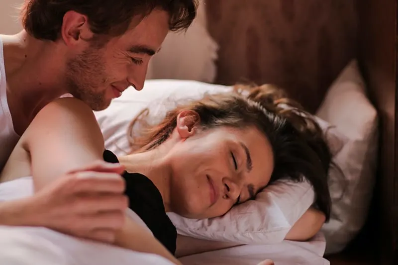 ein lächelnder Mann, der seine schlafende Freundin im Bett ansieht