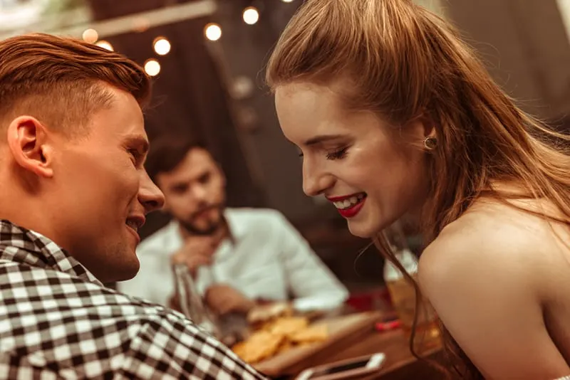 Ein lächelnder Mann und eine lächelnde Frau flirten, während sie mit Freunden im Restaurant sitzen