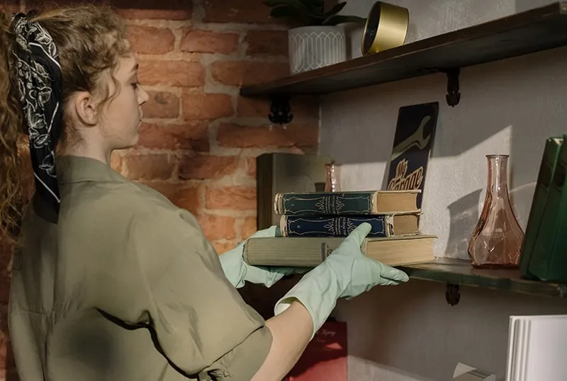 eine Frau, die Bücher in einem Regal organisiert, während sie den Raum putzt