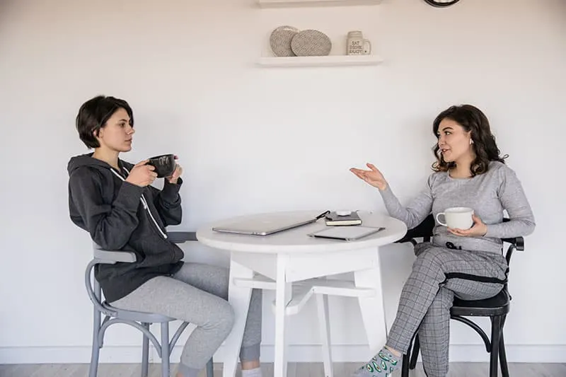 eine ernsthafte Frau, die ihrer lächelnden Freundin zuhört, während sie zusammen Kaffee trinkt