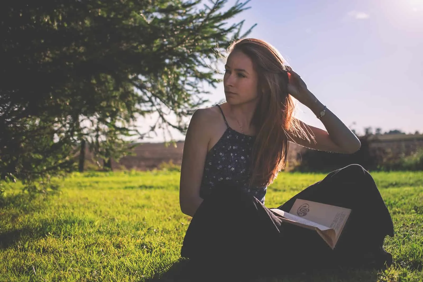 eine nachdenkliche Frau, die beiseite schaut, während sie ein Buch hält und auf dem Gras sitzt