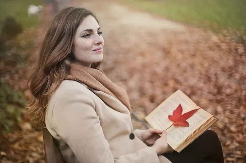 eine positive Frau, die denkt, während sie ein Buch hält und auf der Bank im Park sitzt