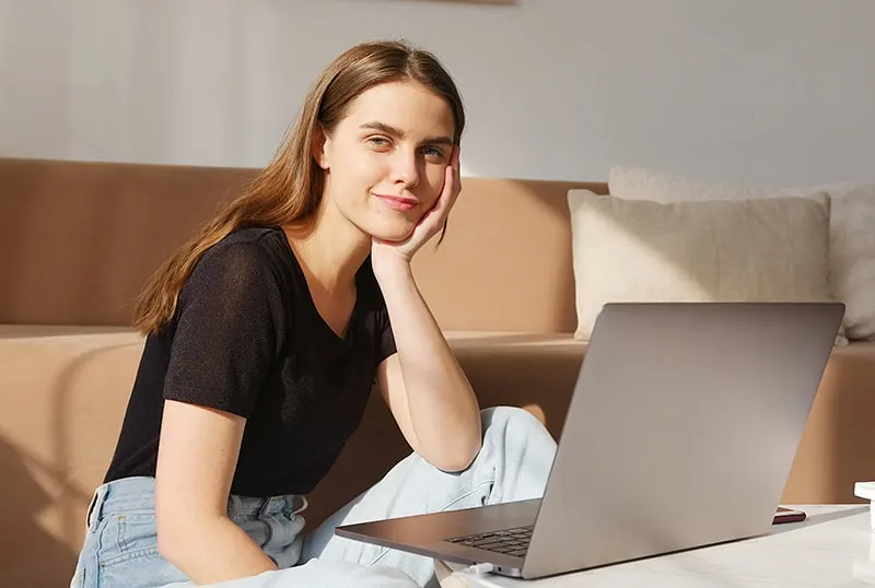 eine positive Frau, die sich auf ihre Hand stützt, während sie vor dem Laptop im Wohnzimmer sitzt