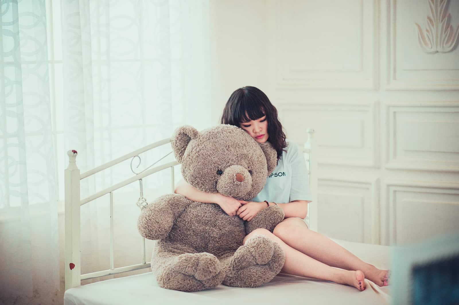 eine traurige Frau, die einen Teddybären umarmt, während sie auf dem Bett sitzt
