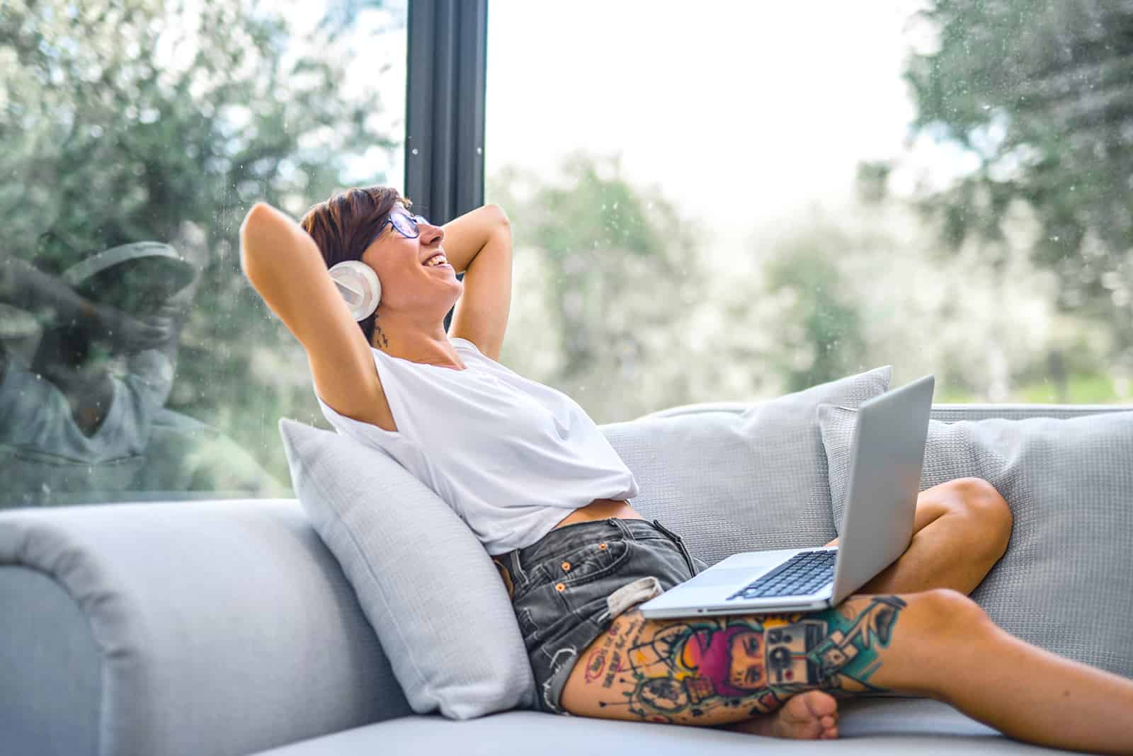 lächelnde Frau mit Kopfhörern, die auf der Couch sitzen, während sie eine Musik auf einem Laptop hören