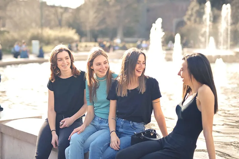 vier lächelnde Freundinnen, die sprechen, während sie auf dem Brunnen sitzen