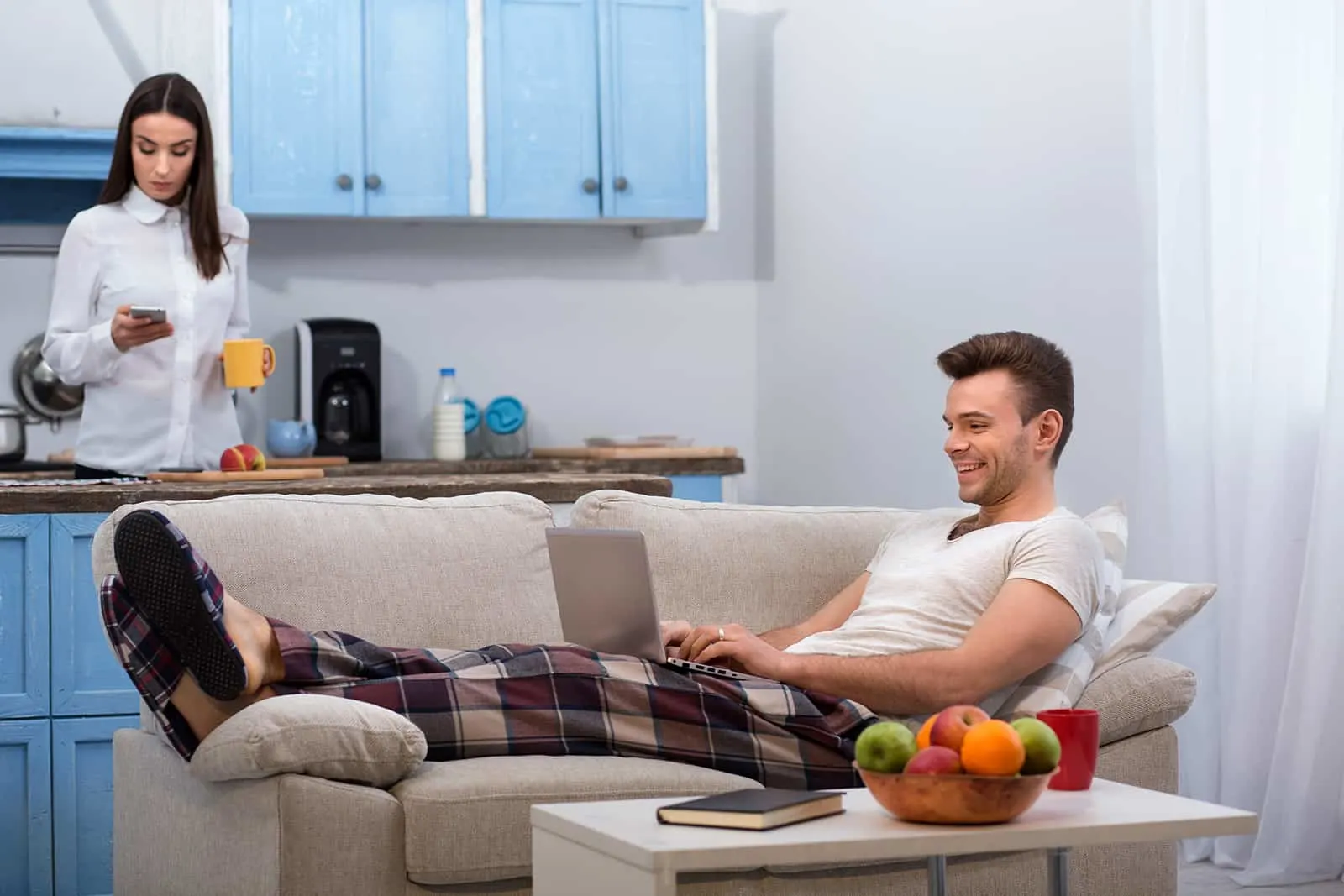 Fauler Ehemann mit einem Laptop auf der Couch, während seine Frau in der Küche steht und sich auf die Arbeit vorbereitet