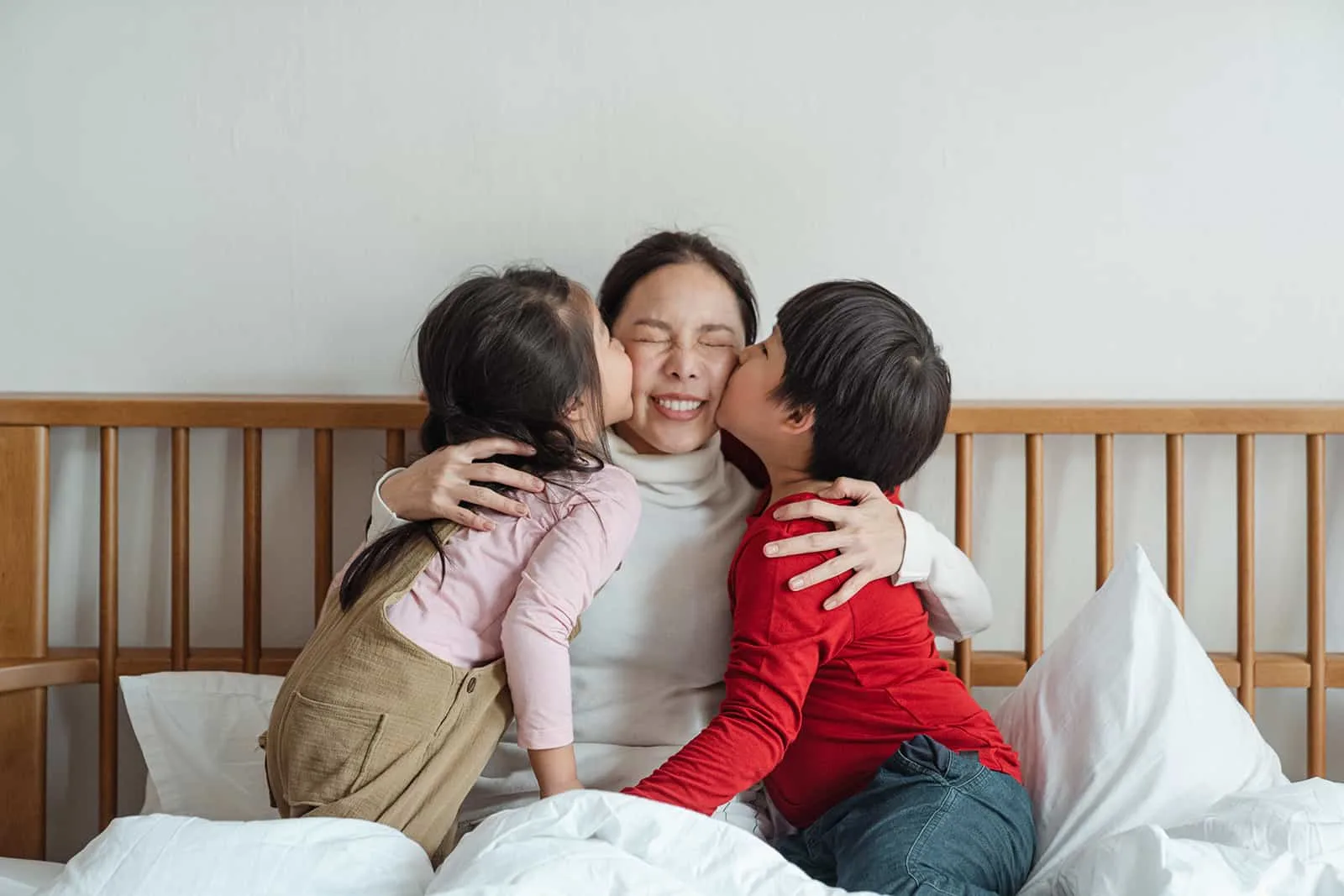 Kinder küssen ihre Mutter auf eine Wange, während sie auf dem Bett sitzen