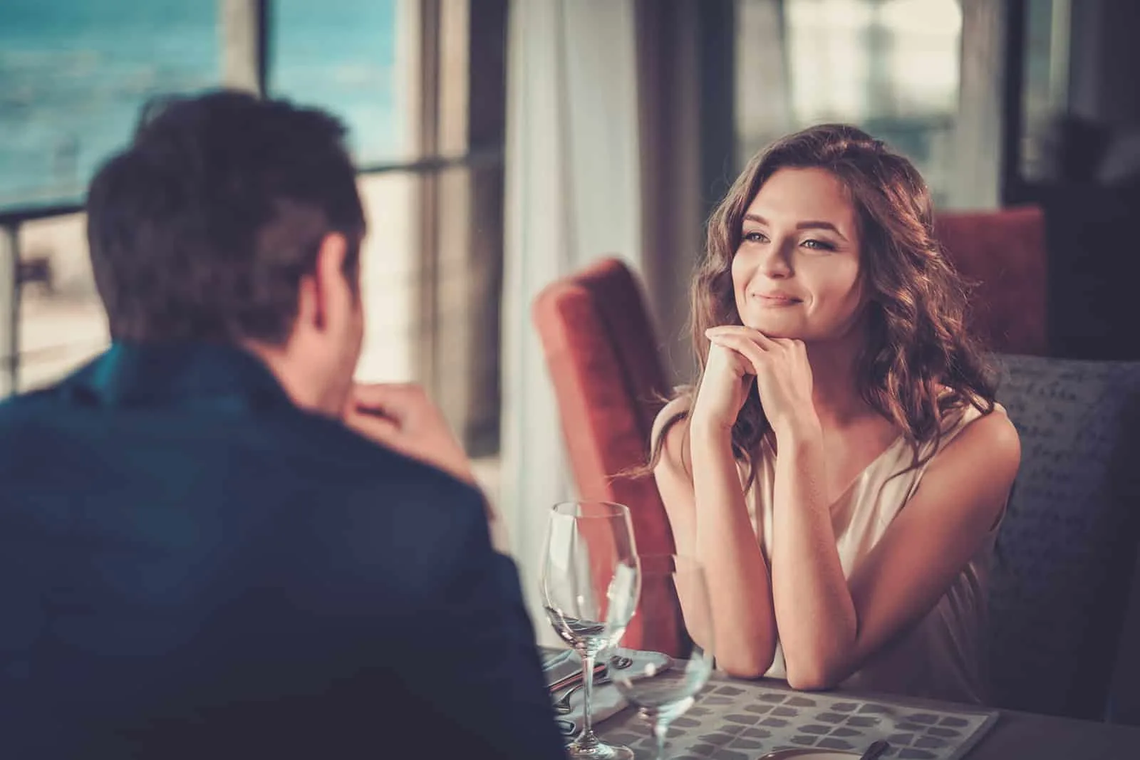 eine lächelnde Frau, die das Kinn auf ihre Hände stützt und auf einen Mann schaut, der mit ihr in einem Restaurant sitzt