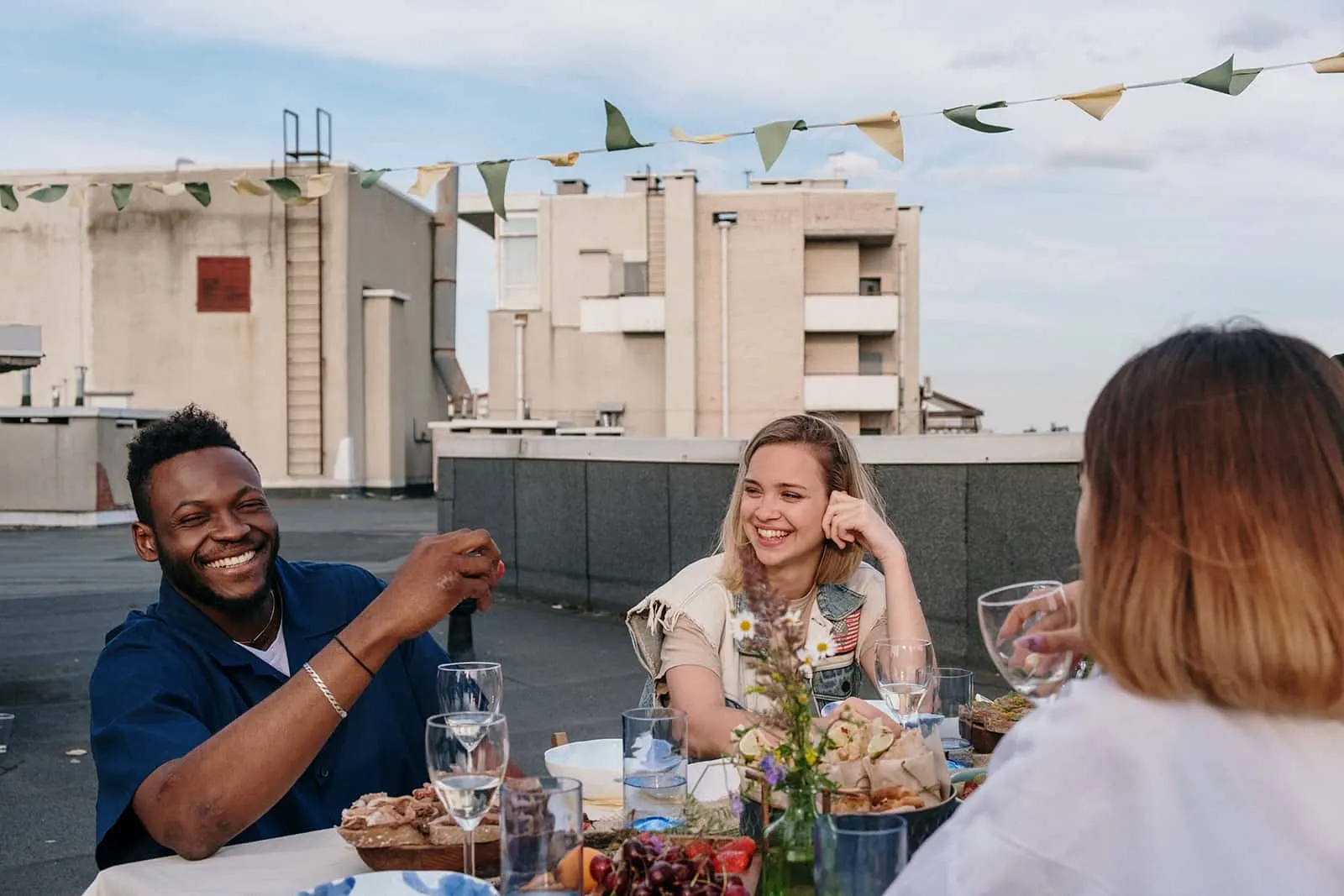 eine lächelnde Frau, die einen lächelnden Mann betrachtet, der mit Freunden auf dem Dach am Tisch sitzt