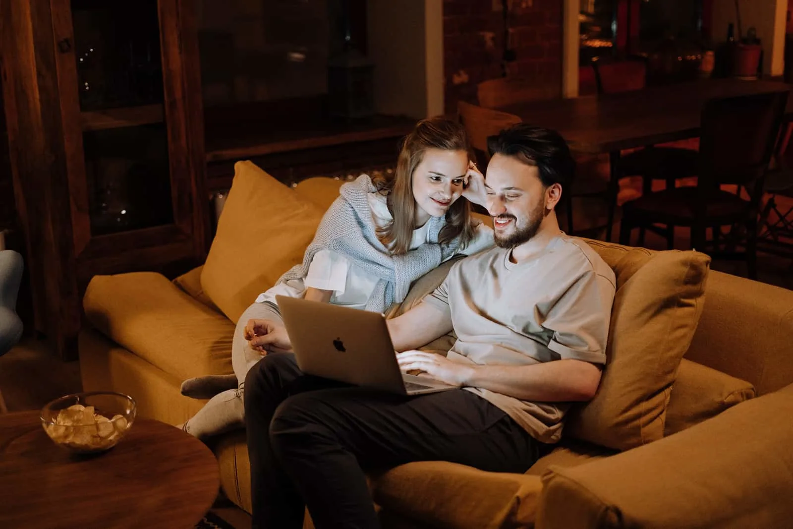 eine lächelnde Frau, die ihren Mann mit einem Laptop betrachtet, der auf der Couch sitzt