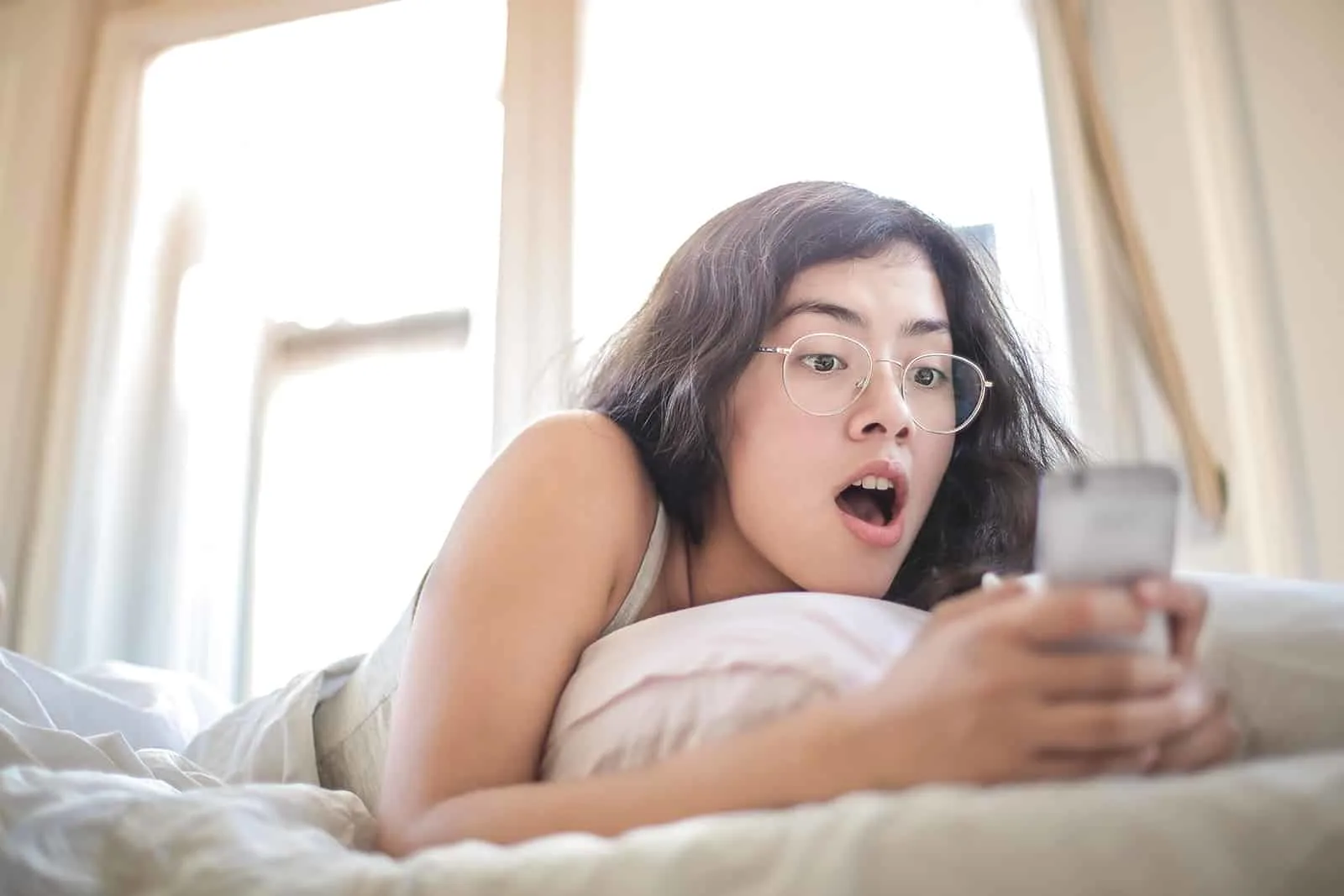 eine überraschte Frau, die ein Smartphone betrachtet, während sie auf dem Bett liegt