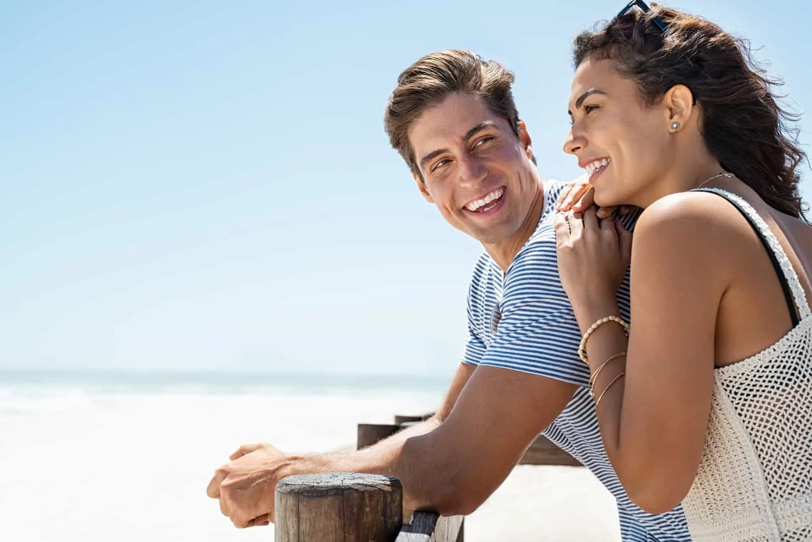 Ein lächelndes Paar, das am Strand steht und sich gegen einen Holzzaun lehnt