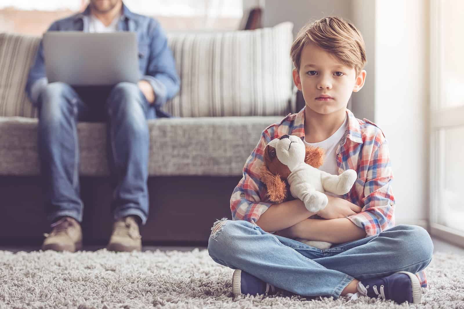 Ein trauriger kleiner Junge sitzt auf dem Teppich, während sein Vater den Laptop auf der Couch benutzt