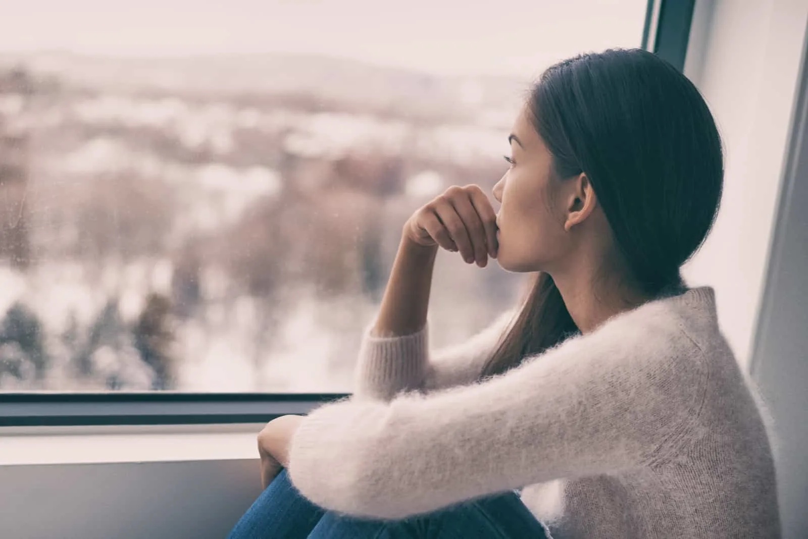 Eine nachdenkliche Frau in einem weißen Pullover sitzt am Fenster und schaut nach draußen