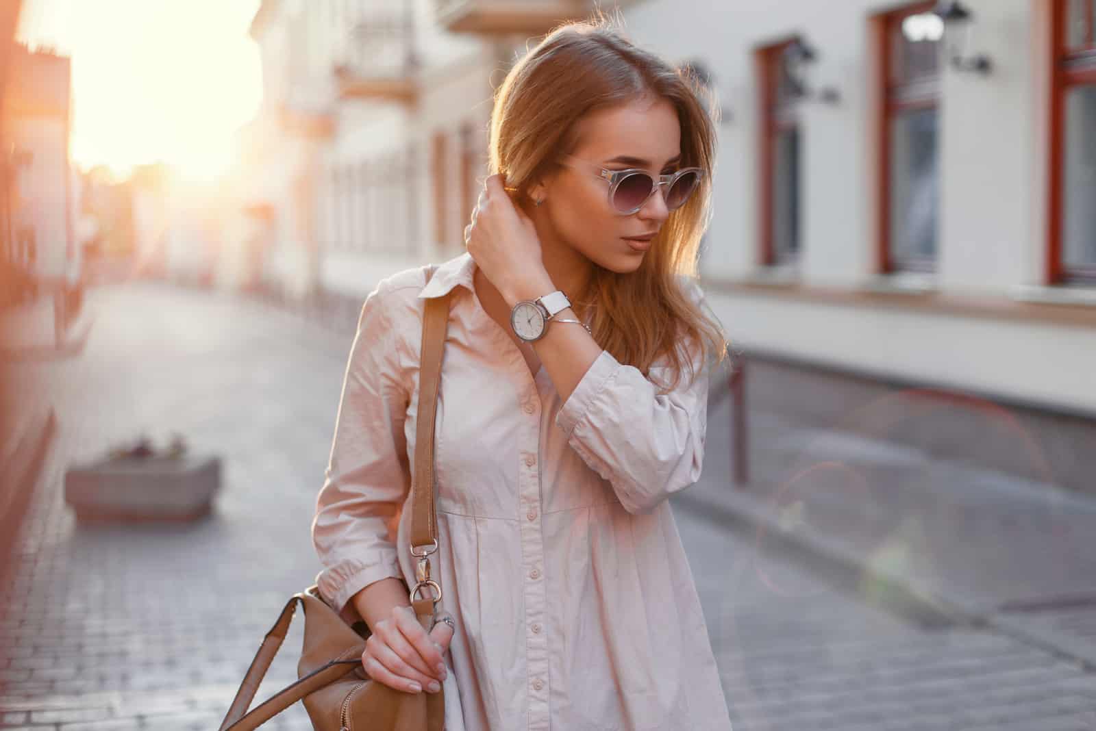 Junges schönes Mädchen in der stilvollen Sonnenbrille und mit einer modischen Tasche bei Sonnenuntergang