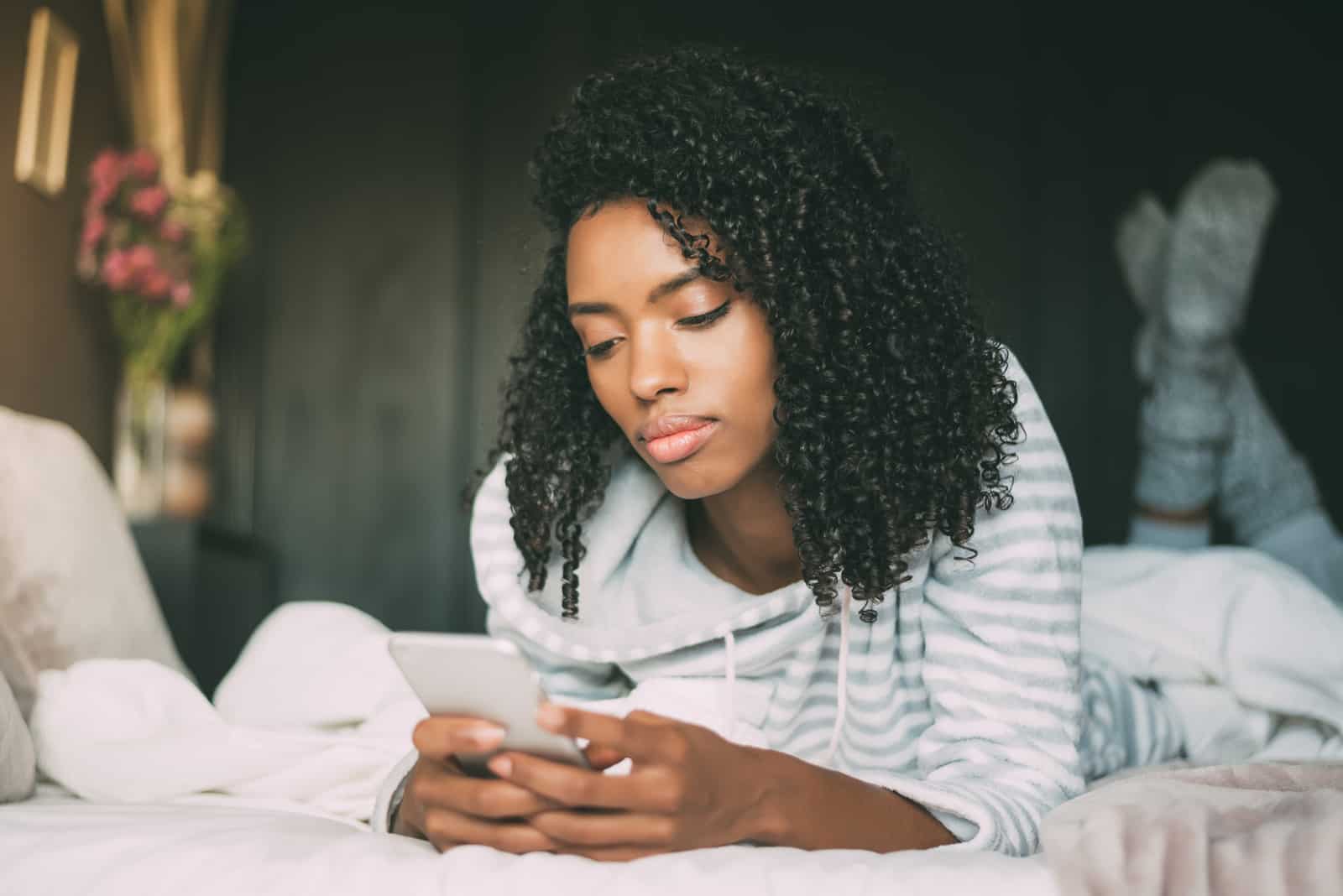 Schöne ernsthafte nachdenkliche und traurige schwarze Frau mit lockigem Haar unter Verwendung des Smartphones auf Bett