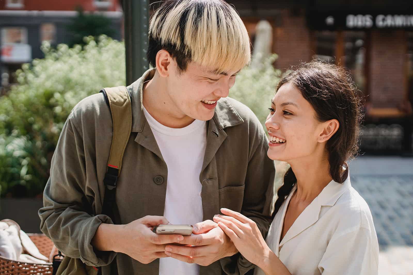 ein lächelnder Mann, der ein Smartphone hält und eine lächelnde Frau betrachtet, die seine Hand berührt