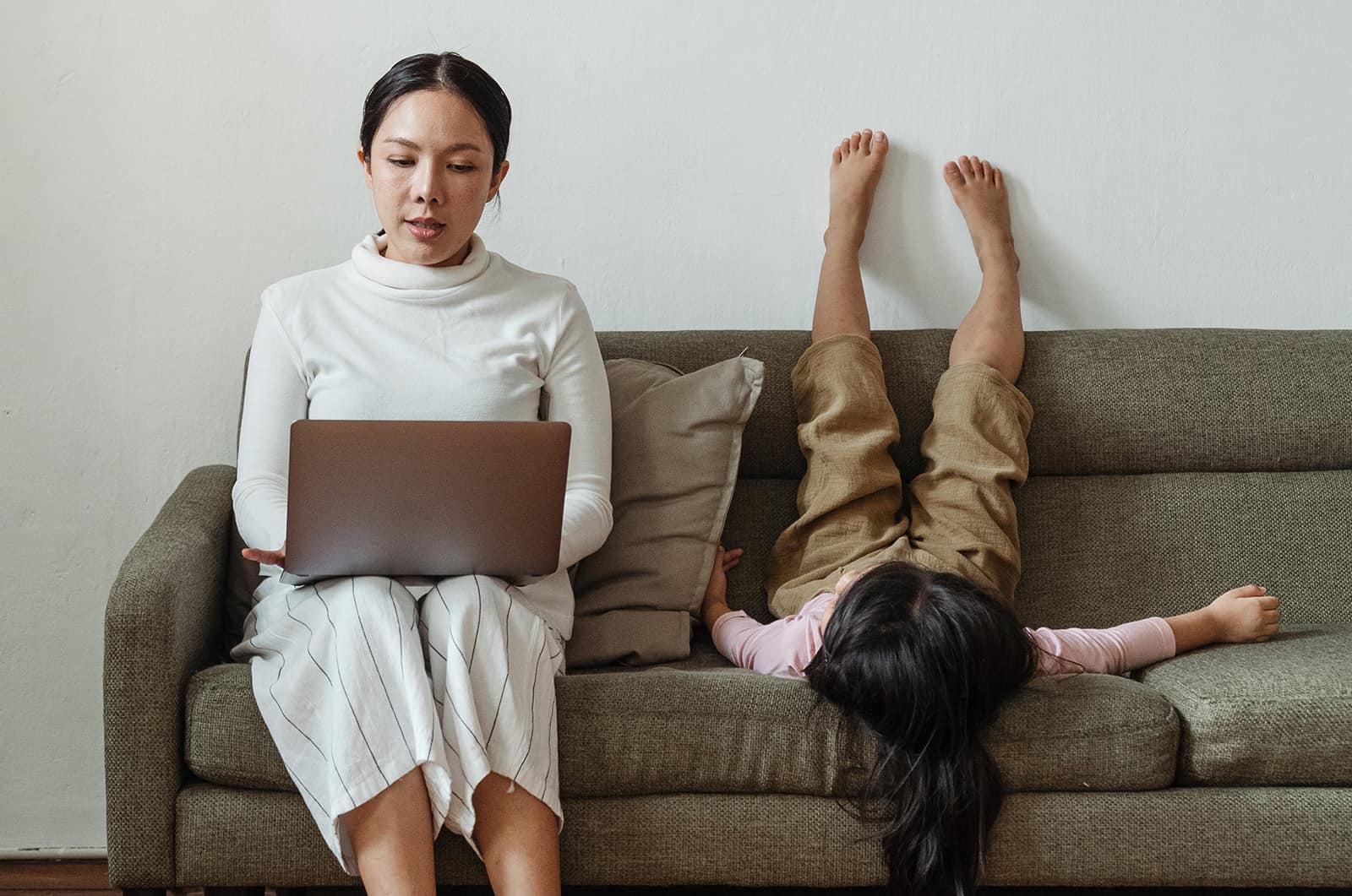 eine fokussierte Mutter, die einen Laptop in der Nähe ihres Kindes benutzt und auf der Couch spielt