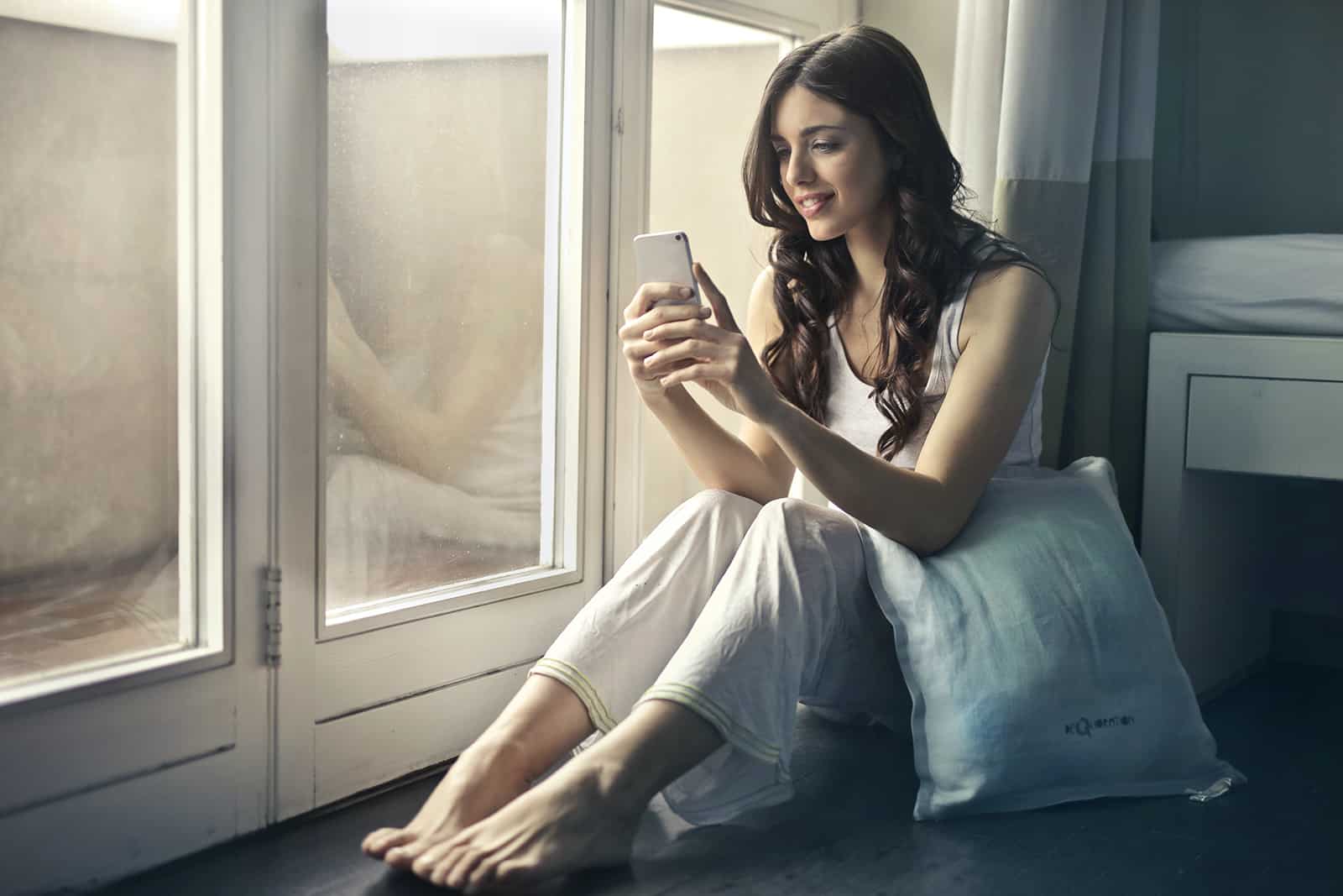 lächelnde Frau, die eine Nachricht auf einem Smartphone tippt, das neben den Glastüren sitzt