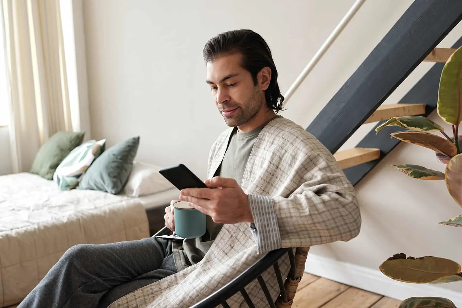 nachdenklicher Mann, der Smartphone beim Sitzen im Sessel zu Hause betrachtet