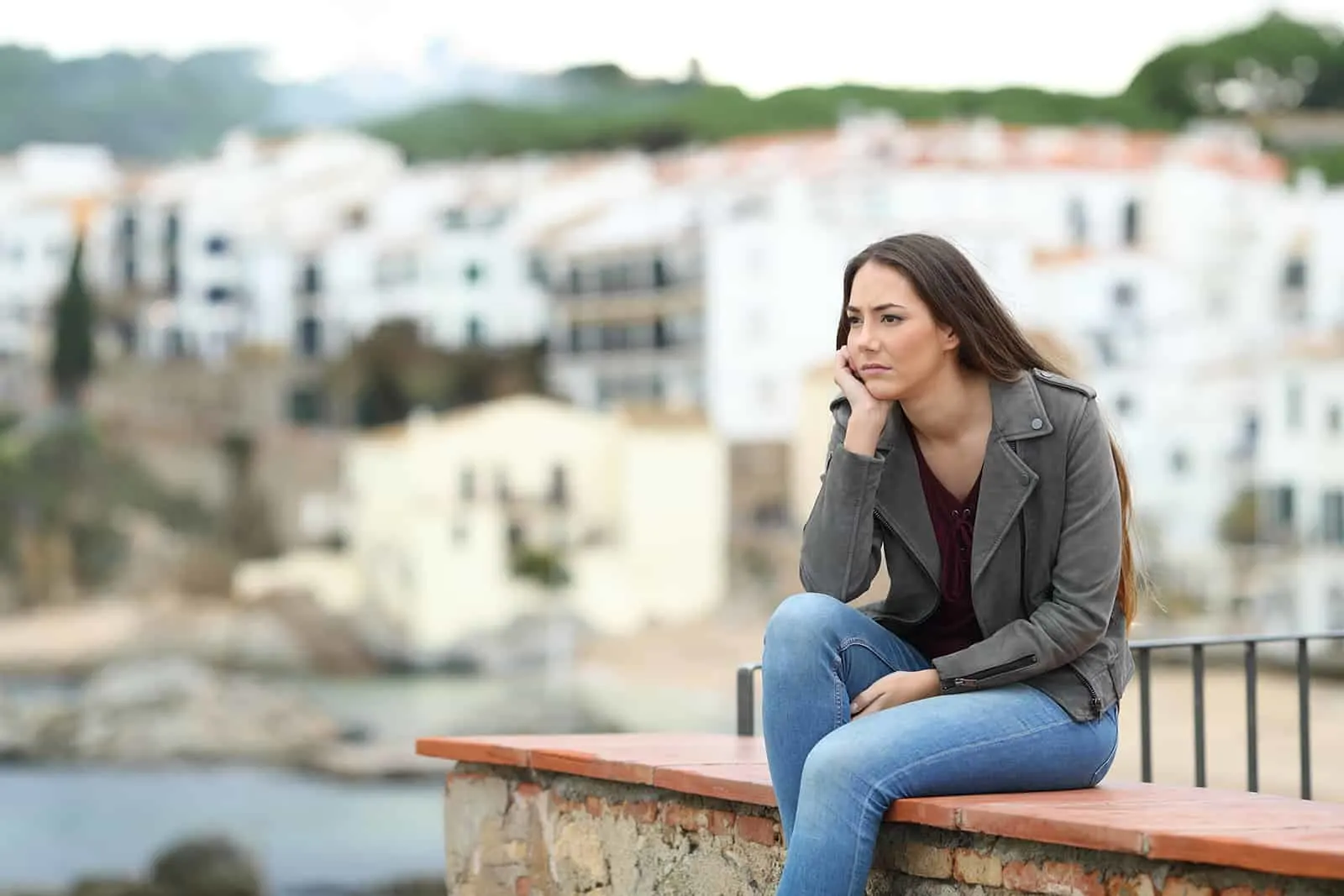 Traurige nachdenkliche Frau, die weg schaut, die auf einem Sims in einer Küstenstadt sitzt