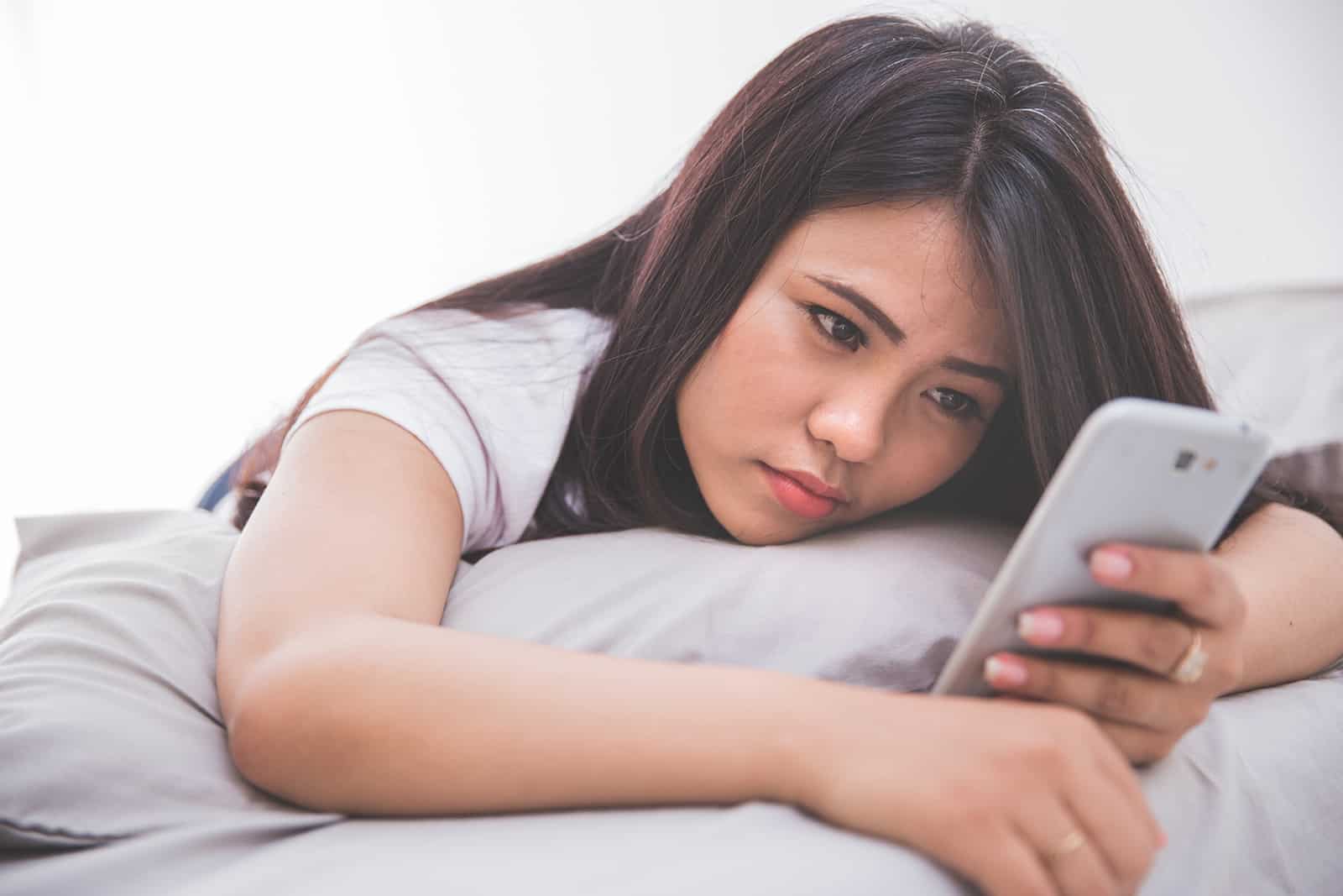 eine Frau, die traurig aussieht und ein Smartphone benutzt, das auf dem Bett liegt