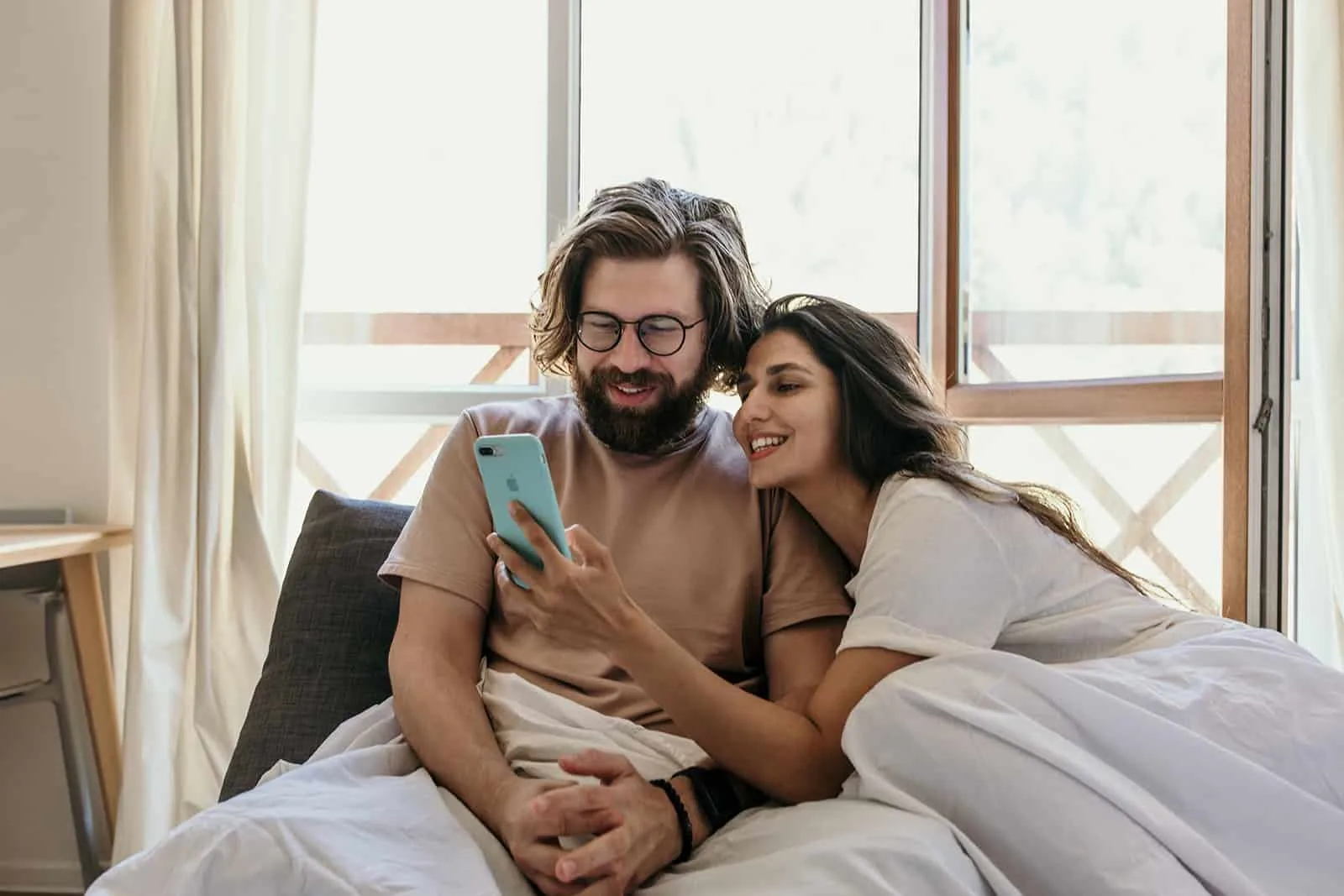 eine lächelnde Frau, die ihrem Freund ein Smartphone zeigt im Bett sitzen