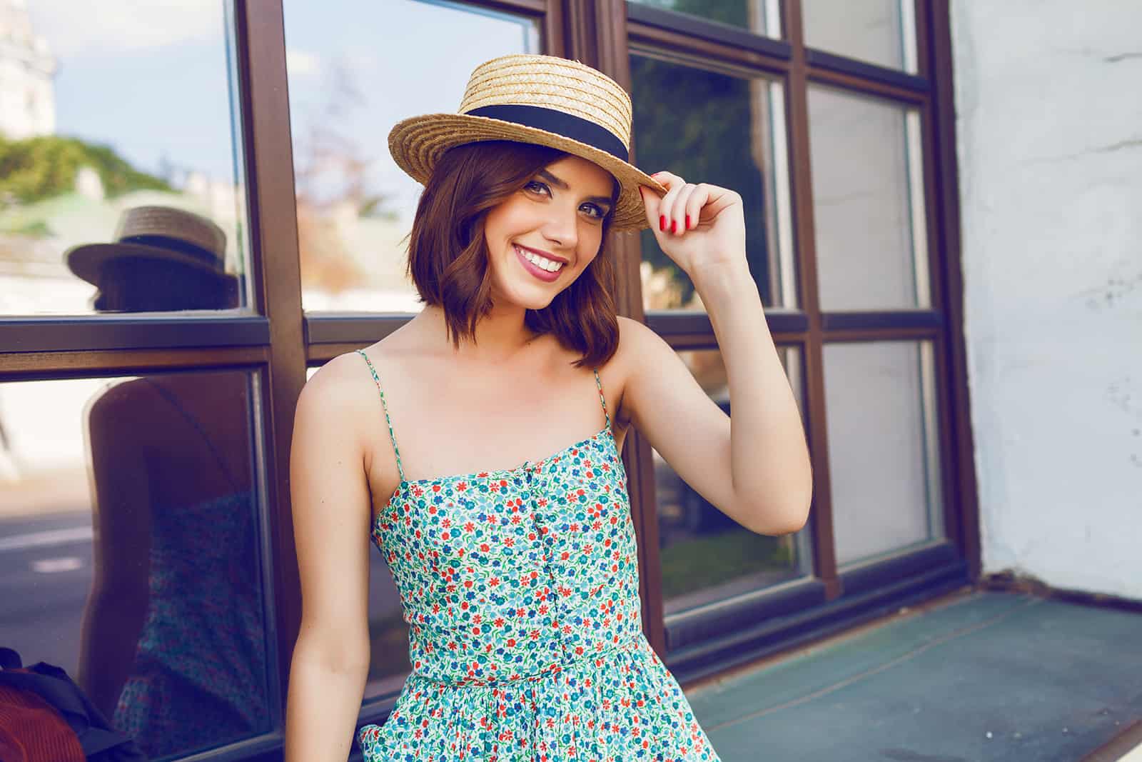 lächelnde Frau in einem Blumenkleid einen Hut mit der Hand berühren