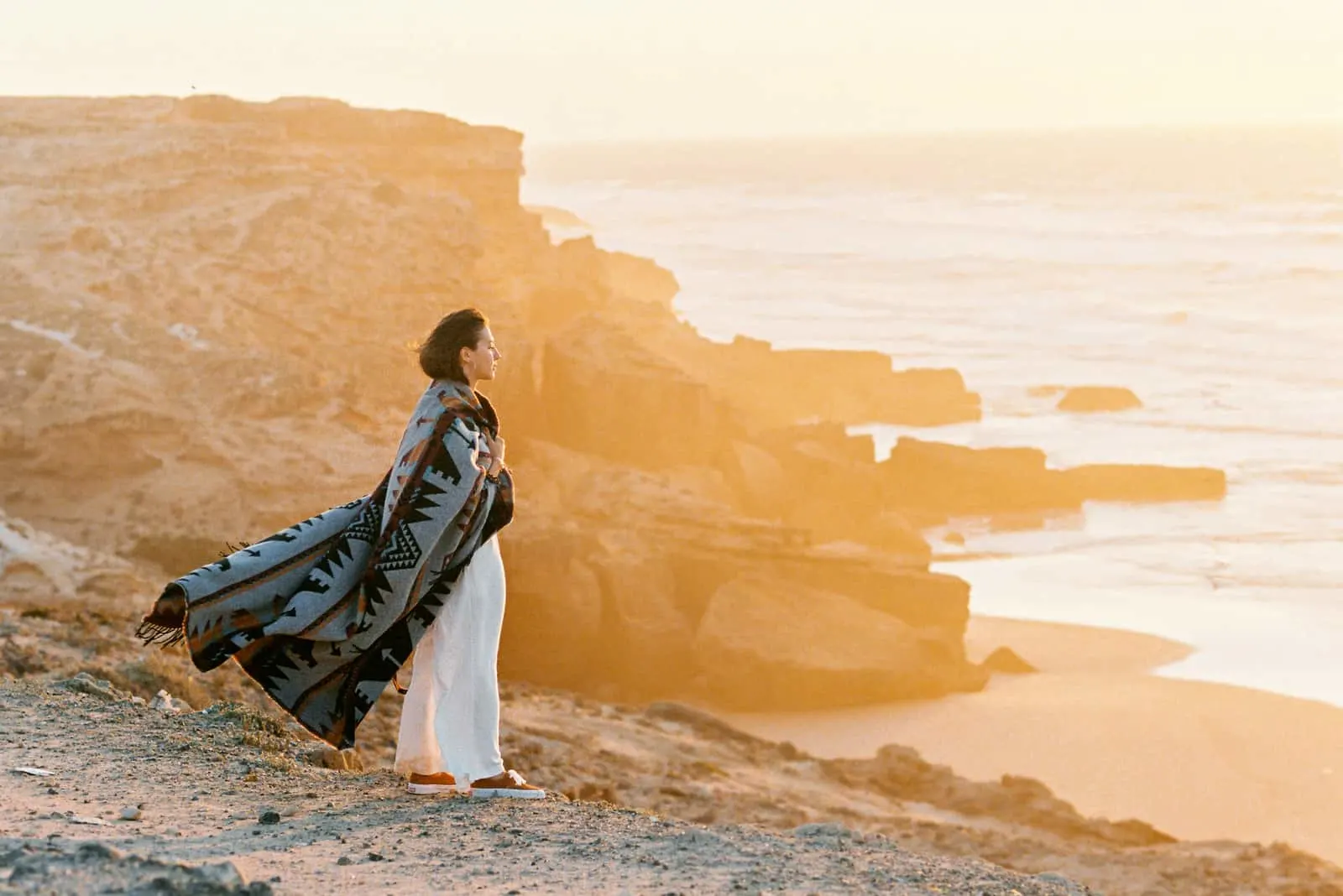 Junge Frau mit fliegender Decke, die auf Klippenrand steht und zum Ozean auf Sonnenuntergang schaut