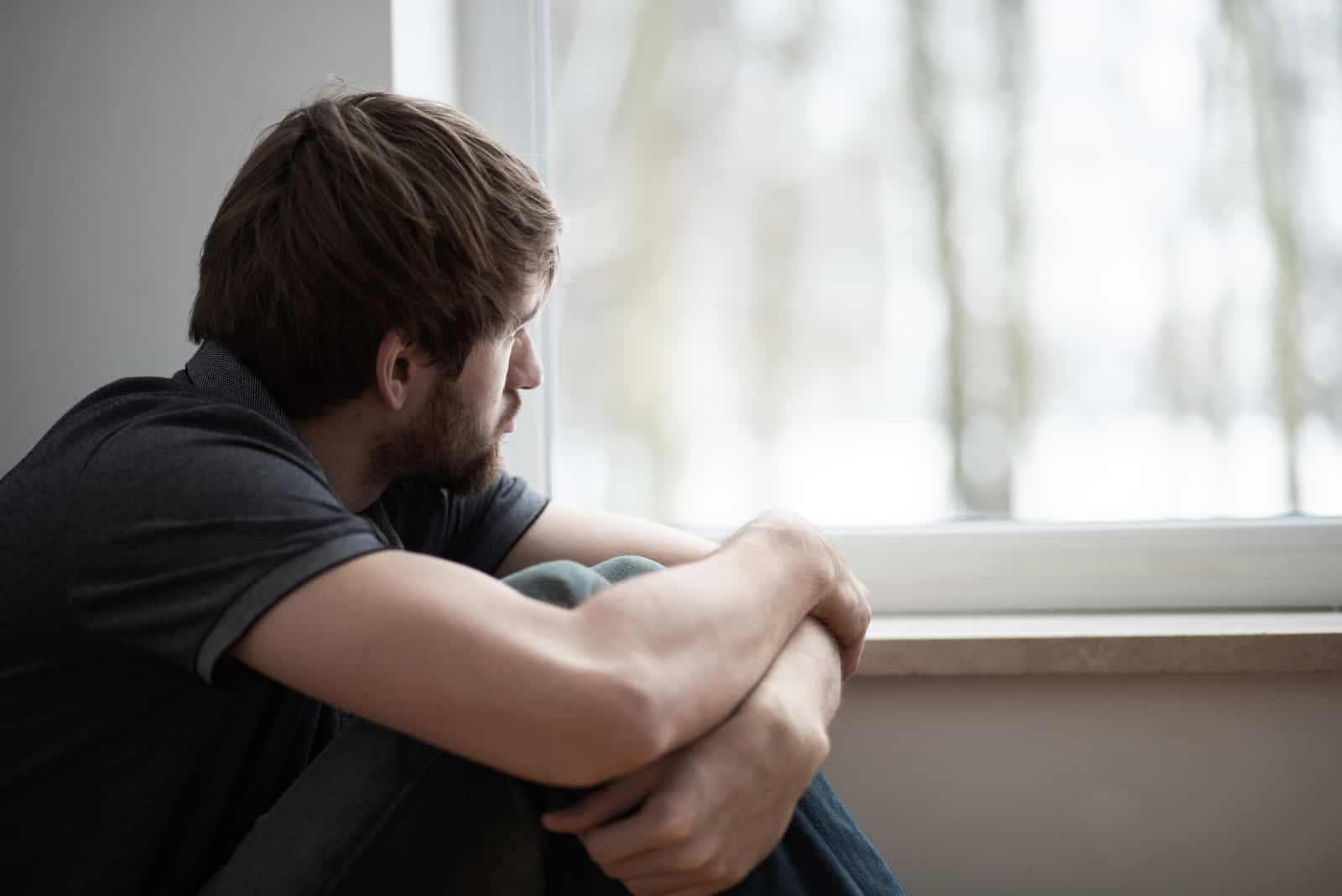 Trauriger junger Mann, der auf dem Boden sitzt und durch das Fenster schaut