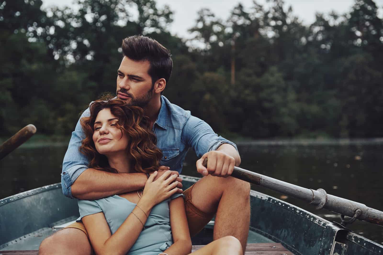 Schönes junges Paar, das sich umarmt und wegschaut, während es ein romantisches Date auf dem See genießt