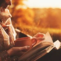 Schönes Mädchen im Herbstwald, das ein Buch liest, bedeckt mit einer warmen Decke, die einen Kaffee hält.