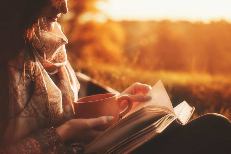 Schönes Mädchen im Herbstwald, das ein Buch liest, bedeckt mit einer warmen Decke, die einen Kaffee hält.