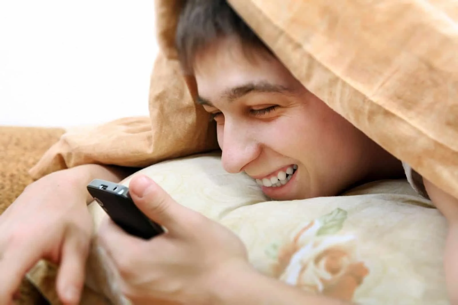 fröhlicher Teenager mit Handy bedeckt mit Decke auf dem Bett liegend