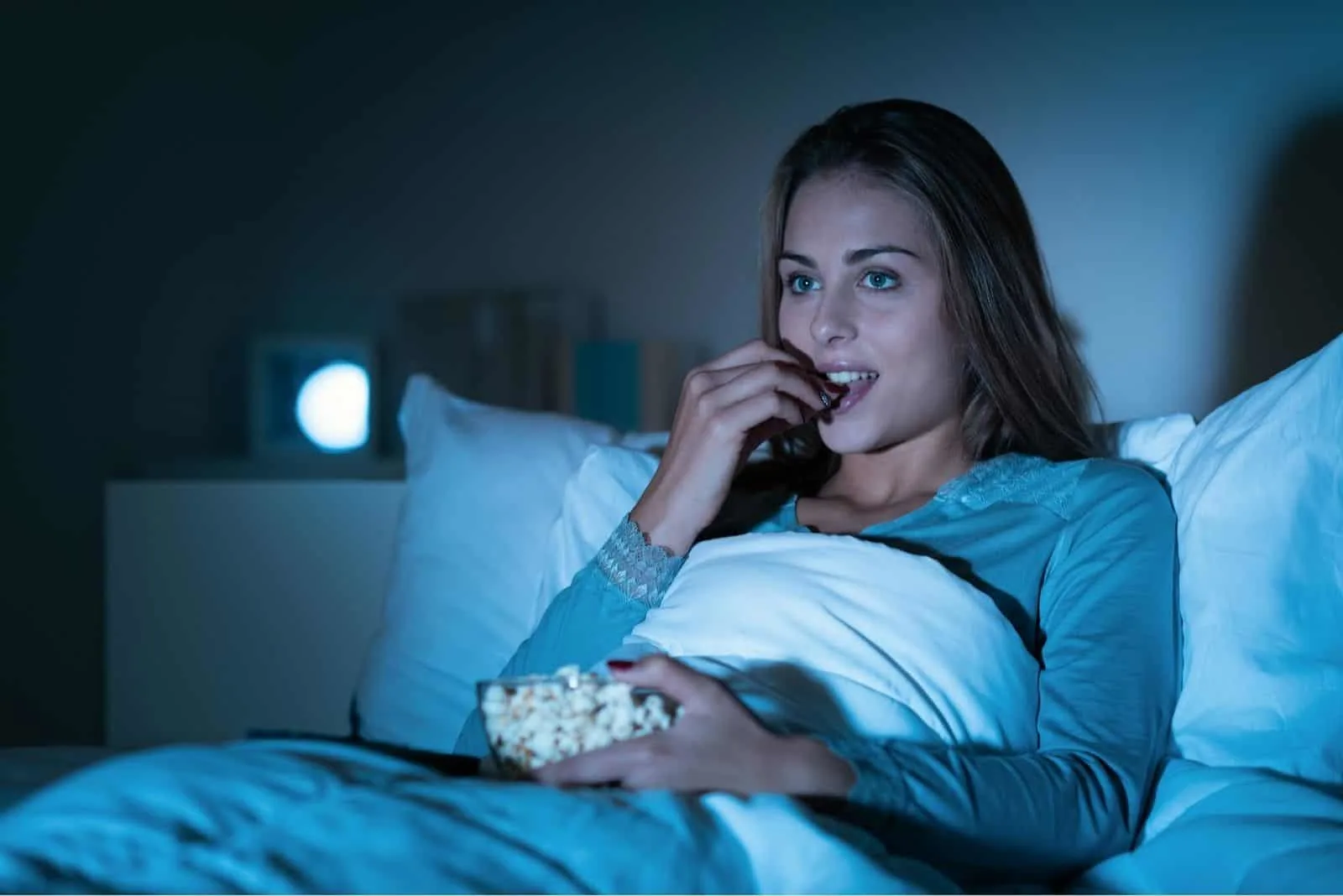 Glückliche Frau, die sich entspannt Nachts im Bett fernsehen und Popcorn essen