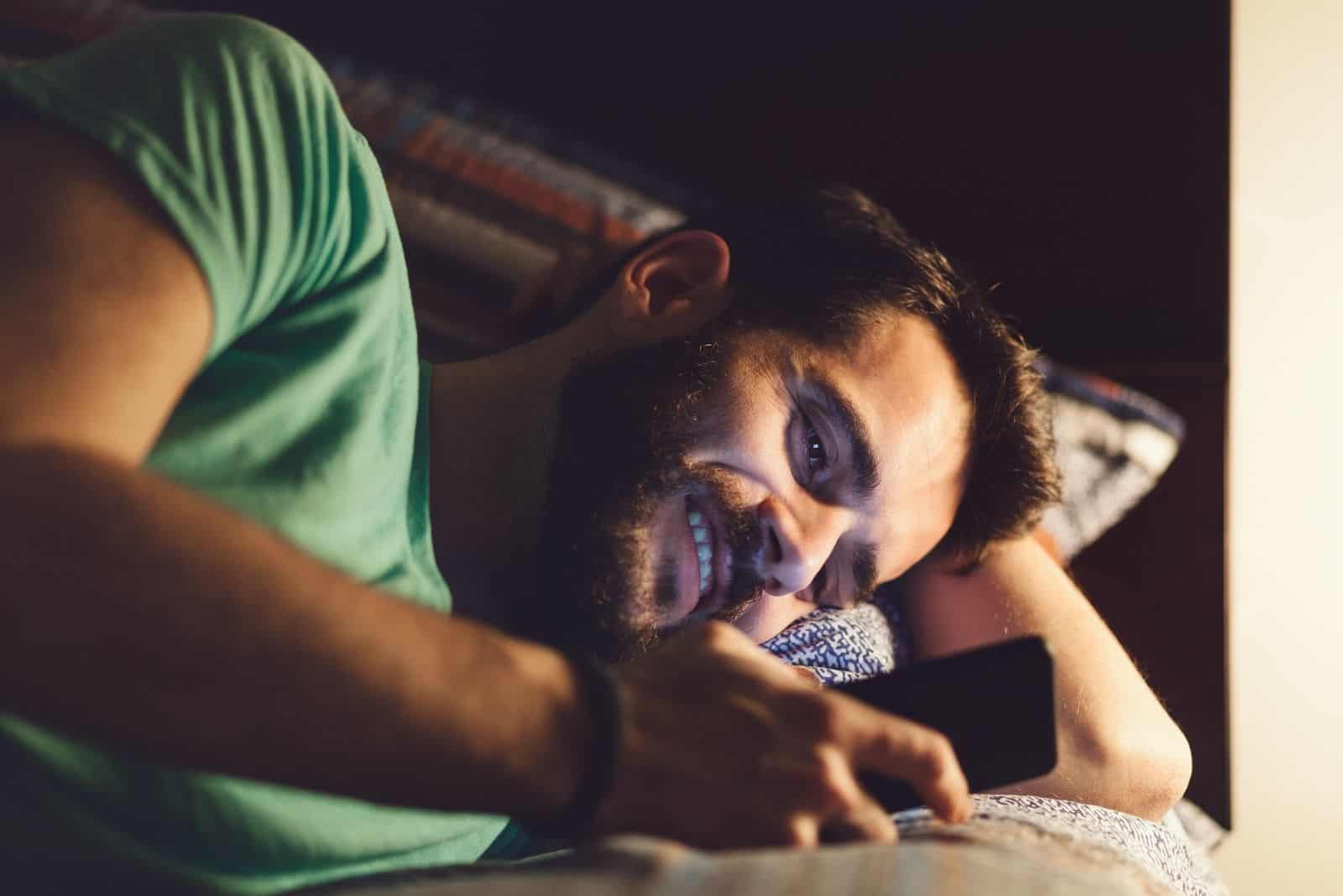 Junger Mann mit einem Smartphone im Bett nachts lächelnd