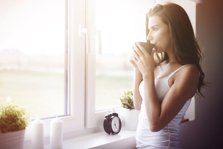 nachdenkliche schöne frau, die morgens kaffee in der nähe der fenster trinkt