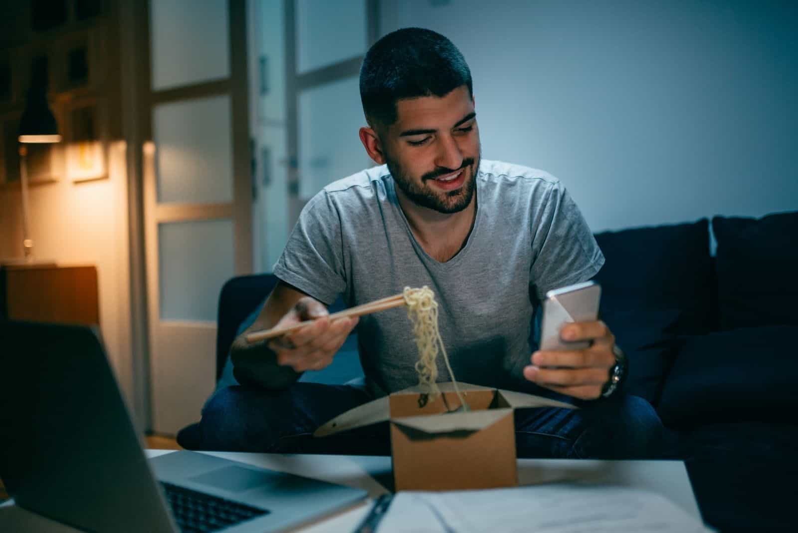 Mann isst chinesisches Essen nachts arbeiten, während er lächelnd auf sein Handy schaut