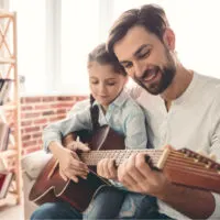 die Tochter sitzt auf dem Schoß ihres Vaters, während sie Gitarre spielen
