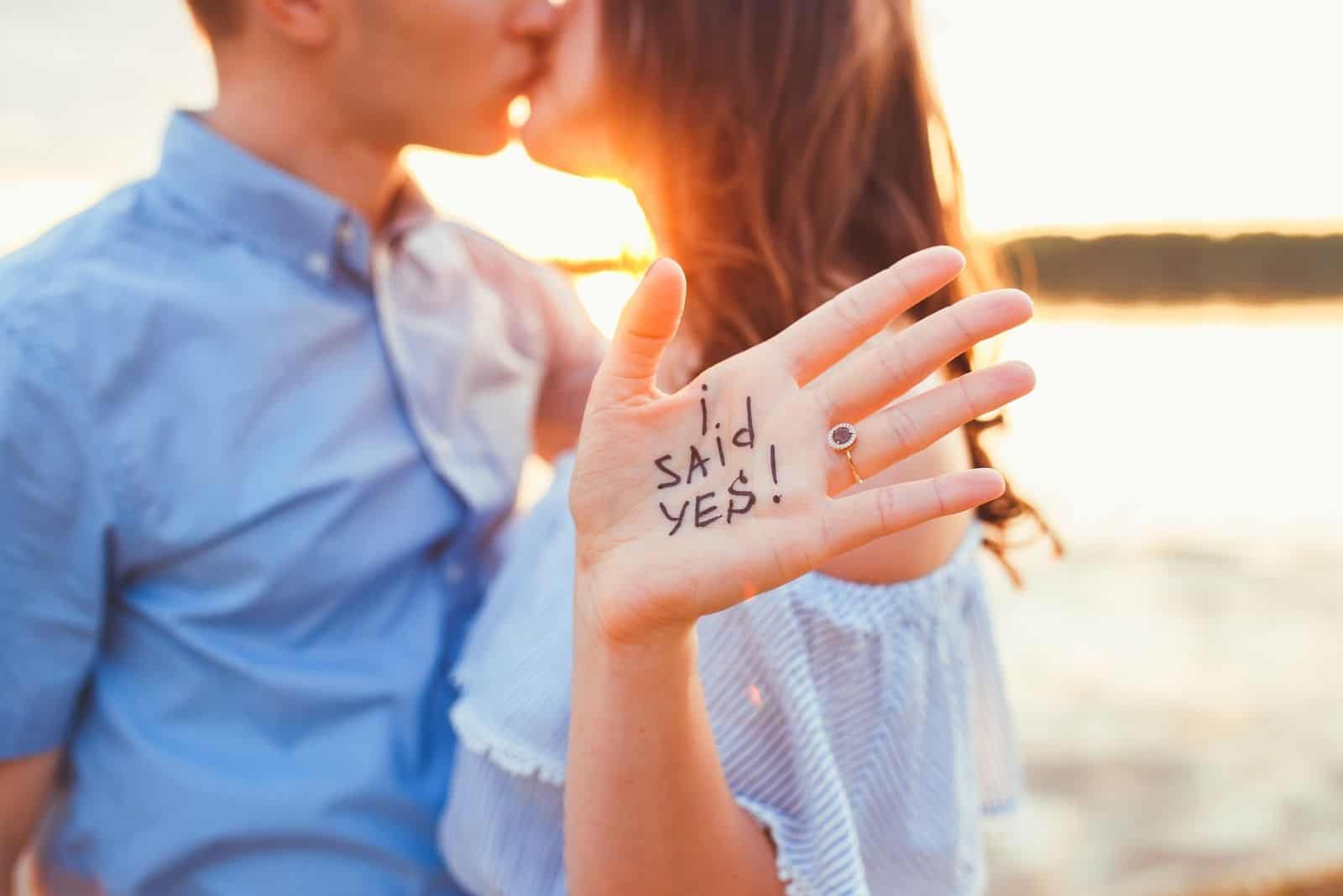 Vorschlag am Strand Frau, die ihre Antwort in der Hand zeigt, während sie sich mit ihrem Freund küsst