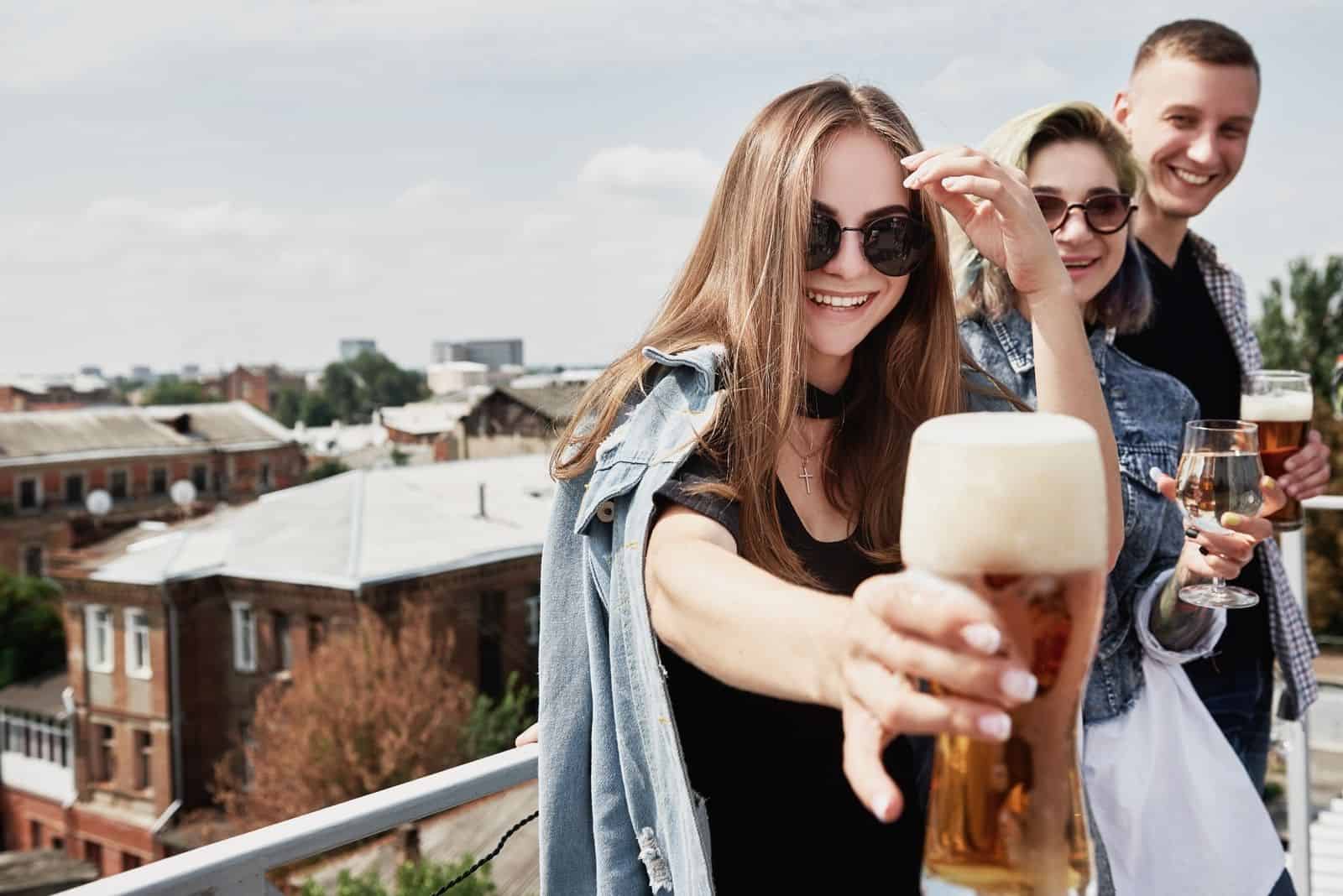 glückliche junge Frau Bier anbieten und dabei Spaß mit Freunden auf dem Dach haben