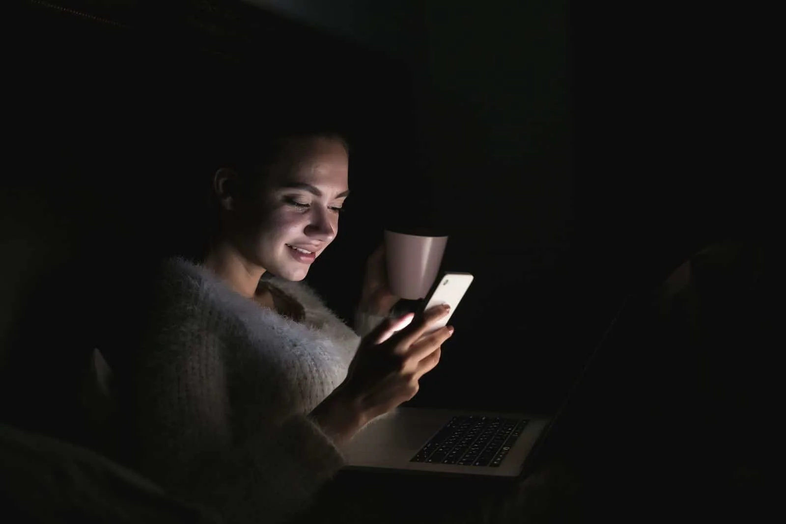 süßes junges Mädchen Nachts im Bett sitzen, mit Laptop, Tee trinken und aufs Telefon schauen, lächeln