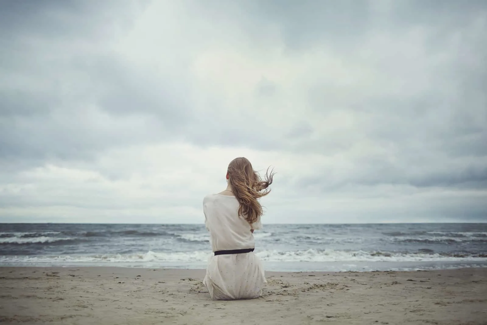 eine Frau mit langen braunen Haaren sitzt am Strand und schaut aufs Meer