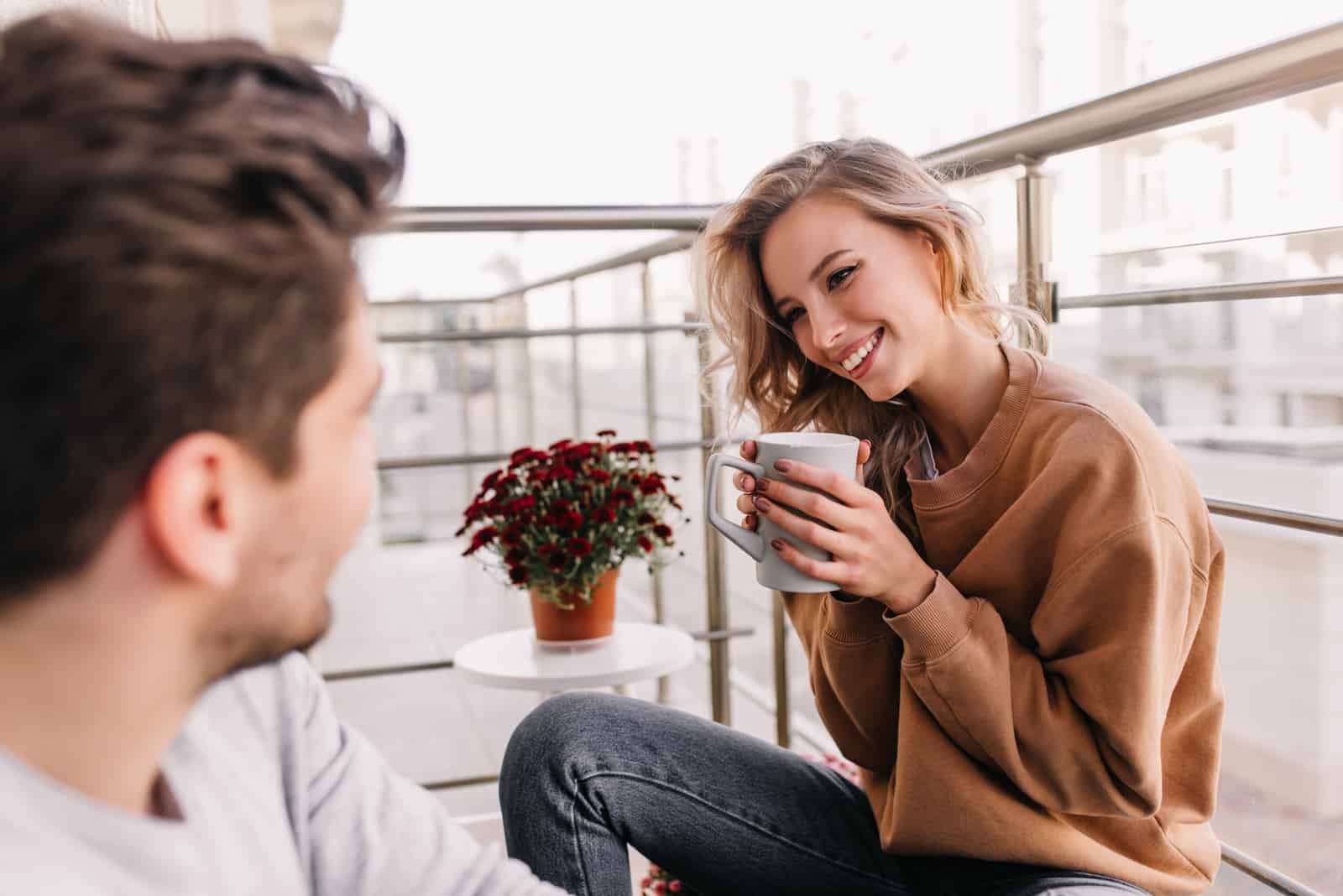 ein lächelnder Mann und eine Frau, die auf dem Balkon sitzen, Kaffee trinken und sich unterhalten