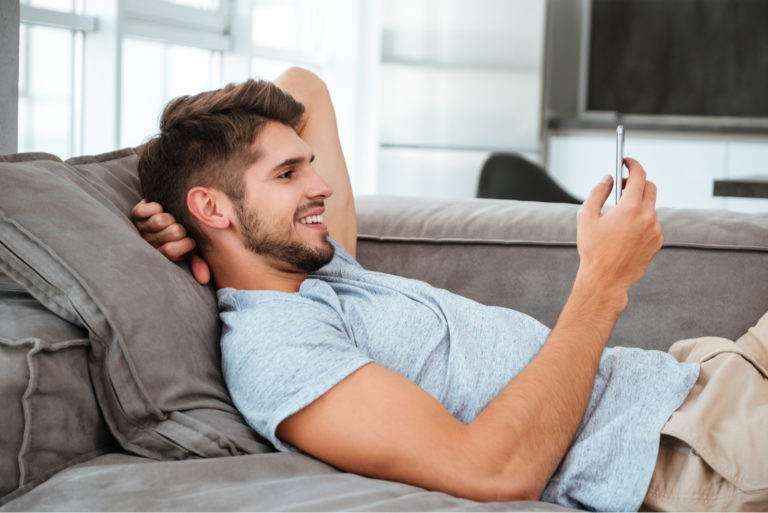 lächelnder gut aussehender Mann, der auf der Couch eine SMS schreibt