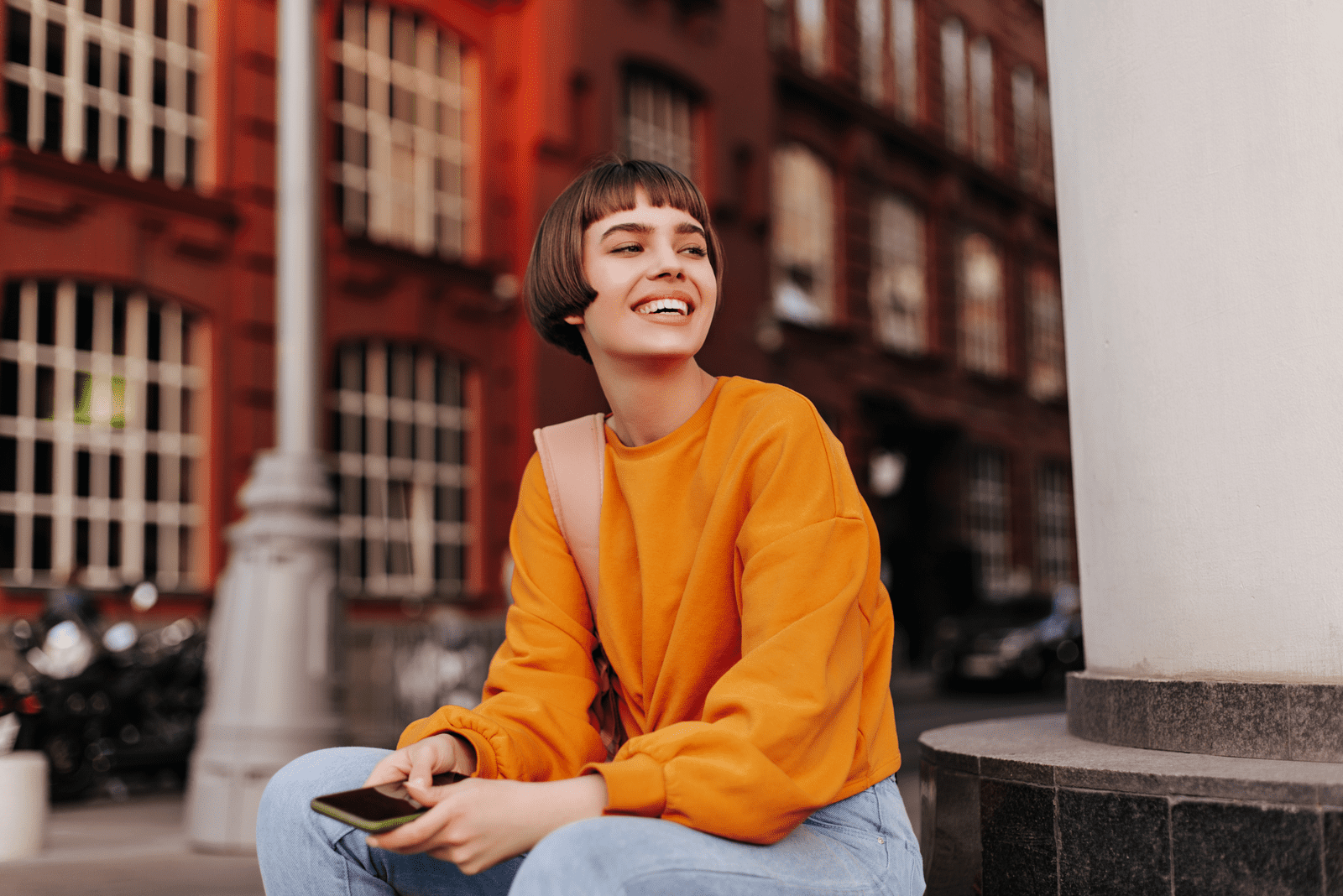 Kurzhaariges Mädchen im orangefarbenen Pullover hält Smartphone und lächelt im Freien