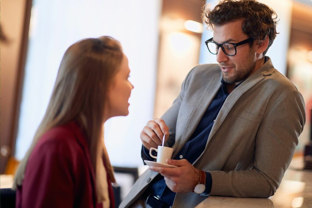 Ein Mann lehnt an der Bar, ruehrt Kaffee um und spricht mit einer Frau