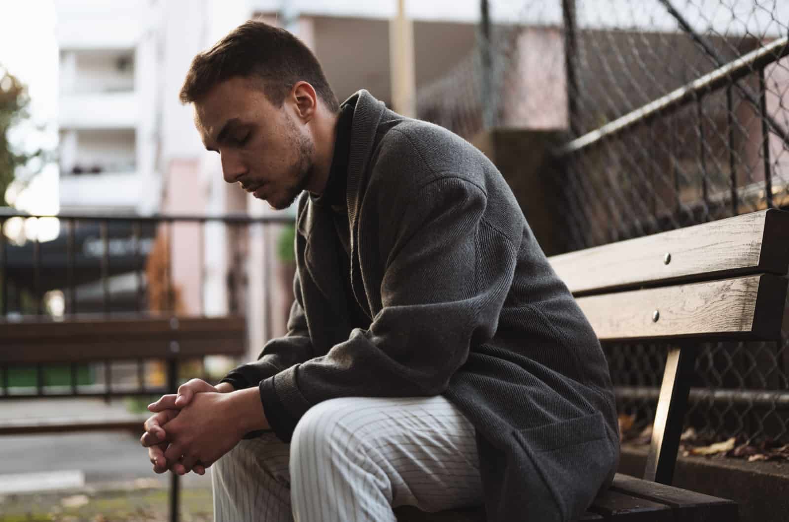 Junger Mann im dunklen Mantel sitzt draußen auf einer Holzbank